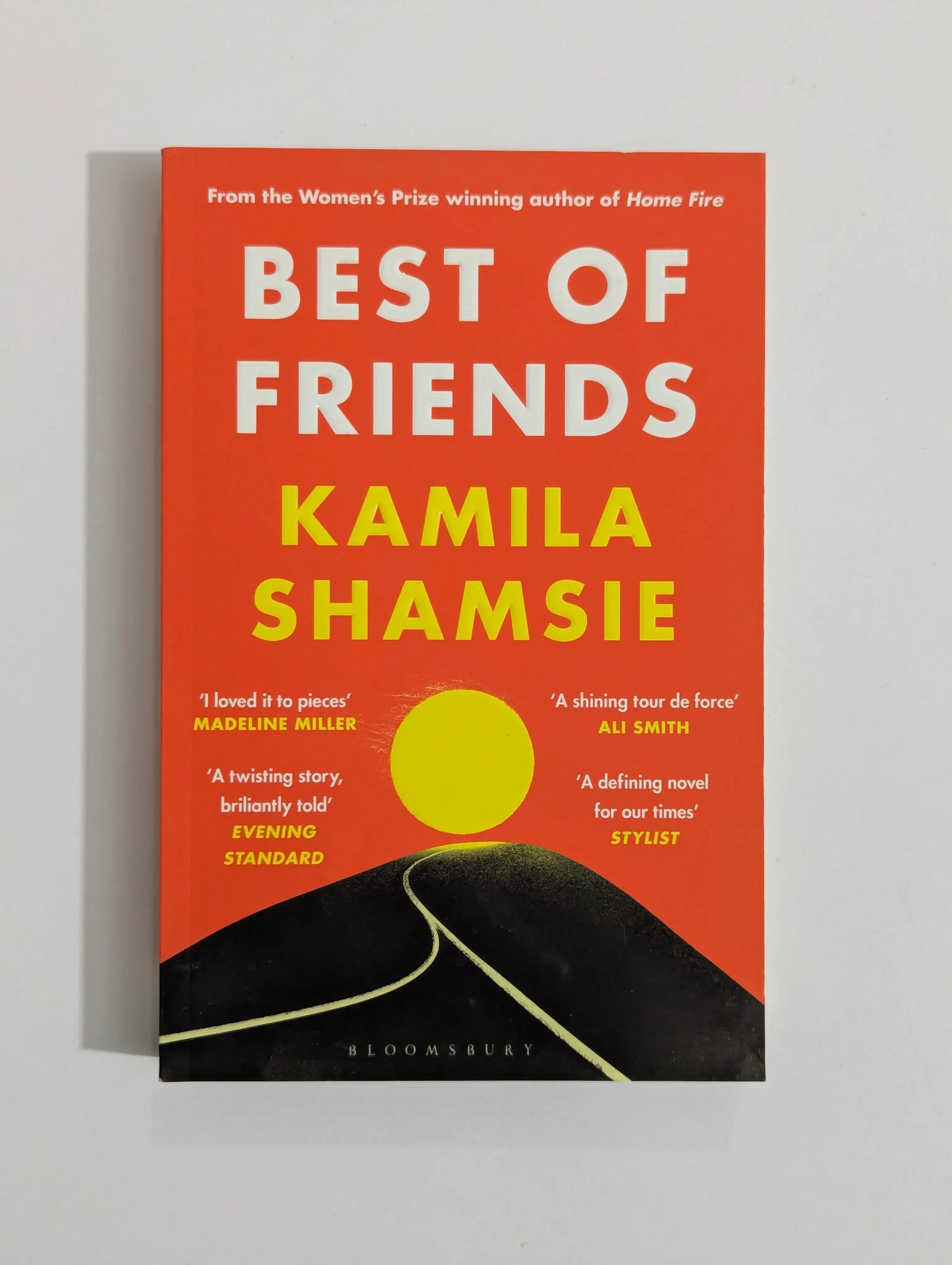 The book cover of 'Best of Friends' by Kamila Shamsie, featuring a bold orange-red background with large white and yellow title text, a yellow circle graphic, and a black road illustration at the bottom. The Bloomsbury publisher name is visible at the bottom.