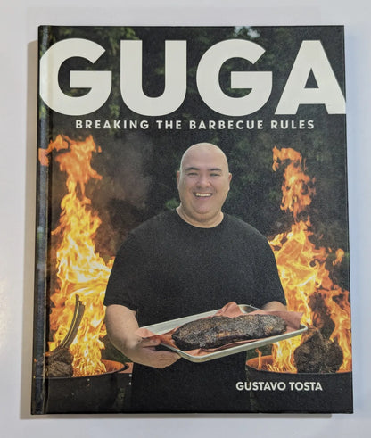 Book titled 'Guga: Breaking the Barbecue Rules' by Gustavo Tosta with a person holding a roasted item in front of flames on the cover.