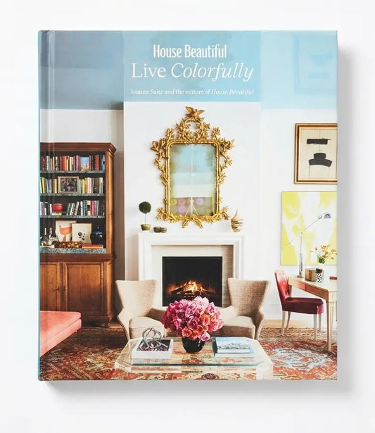 The book 'House Beautiful Live Colorfully' by Susan Succi is shown standing upright against a clean white background. The cover features a colorful photograph of a beautifully decorated living room with a fireplace, gold mirror, bookshelves, and floral arrangements.