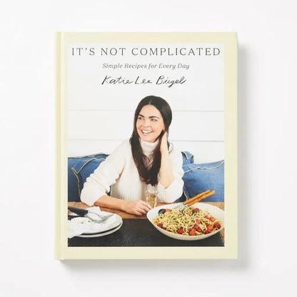 The front cover of the cookbook 'It's Not Complicated: Simple Recipes for Every Day' by Katie Lee Diegel, featuring a cream-colored border and a photo of a smiling woman with a bowl of pasta on a table.