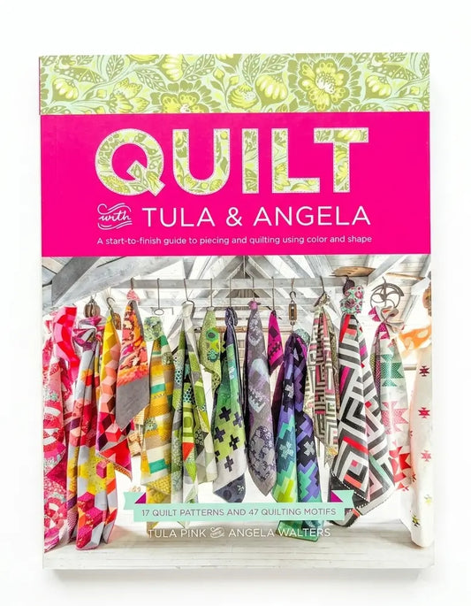 Quilt Tula & Angela book cover featuring a bright pink title band and a photo of colorful quilted fabrics hanging on a rack.