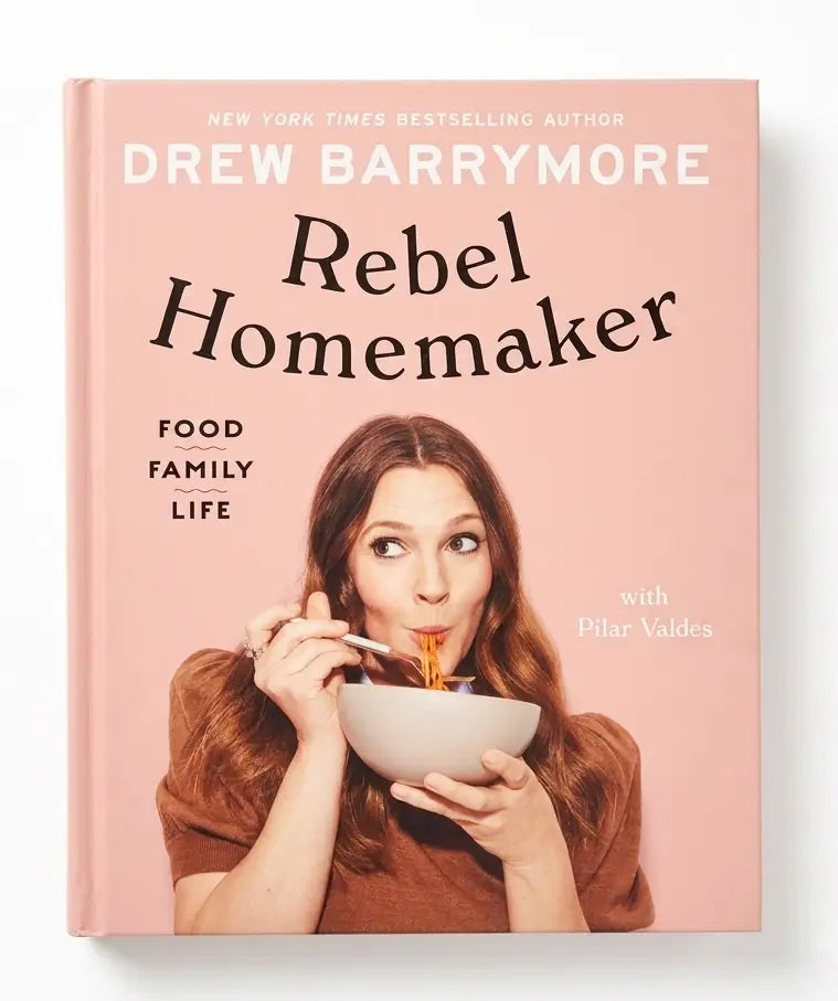 The book cover of 'Rebel Homemaker' by Drew Barrymore, featuring a pink background with the author's name and title in large text, and a photo of Drew Barrymore eating pasta from a bowl.