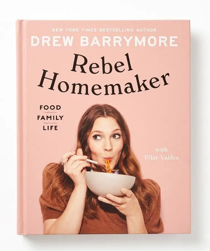 The book cover of 'Rebel Homemaker' by Drew Barrymore, featuring a pink background with the author's name and title in large text, and a photo of Drew Barrymore eating pasta from a bowl.