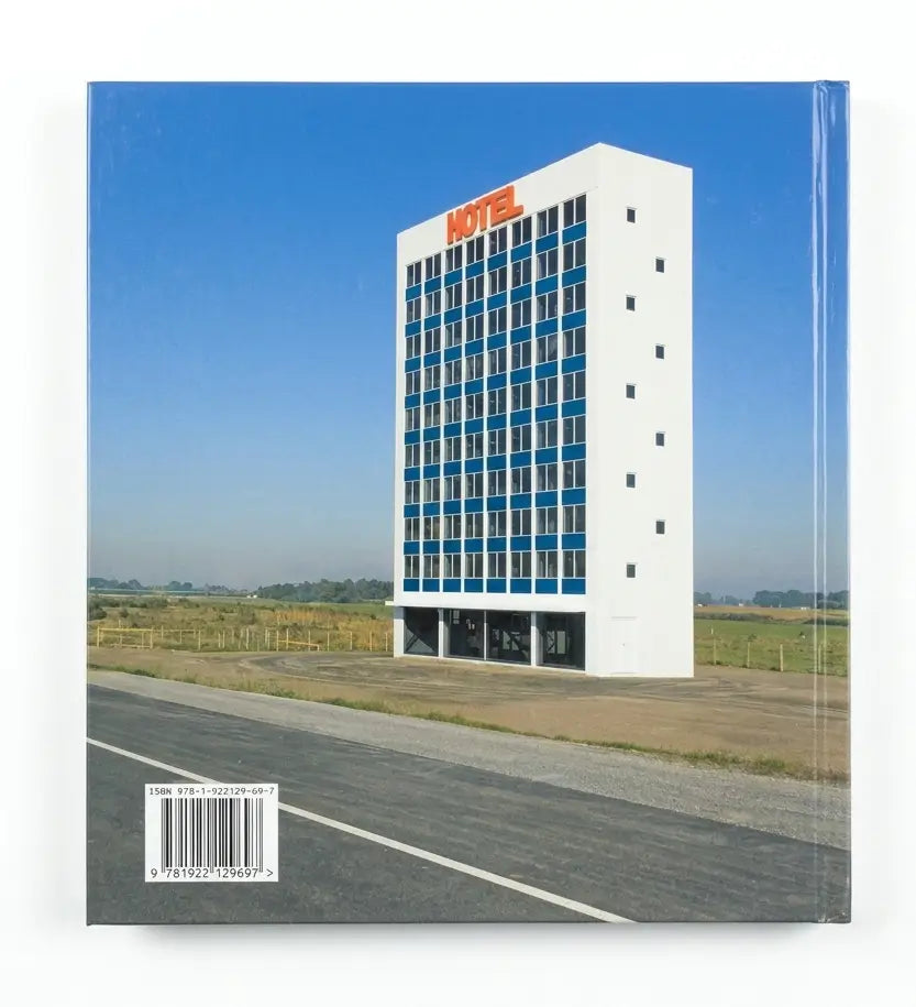A square art print or book cover featuring a surreal photograph of a tall white motel building with blue-tinted windows beside a road under a clear blue sky. The word 'MOTEL' appears in red on the roof, and a barcode is visible in the lower left corner.