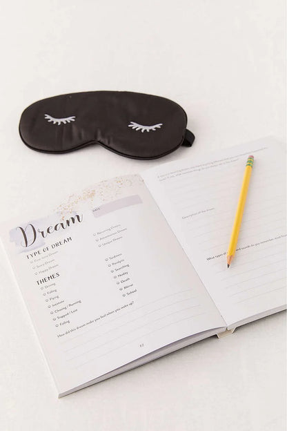 Sleep mask with eyelashes next to an open notebook with a pencil on a white background
