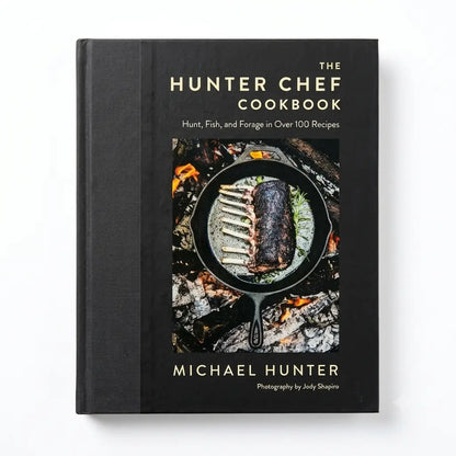 The Hunter Chef Cookbook by Michael Hunter, a hardcover book with a black cover and gold lettering, featuring a central image of ribs cooking in a cast iron skillet over embers.