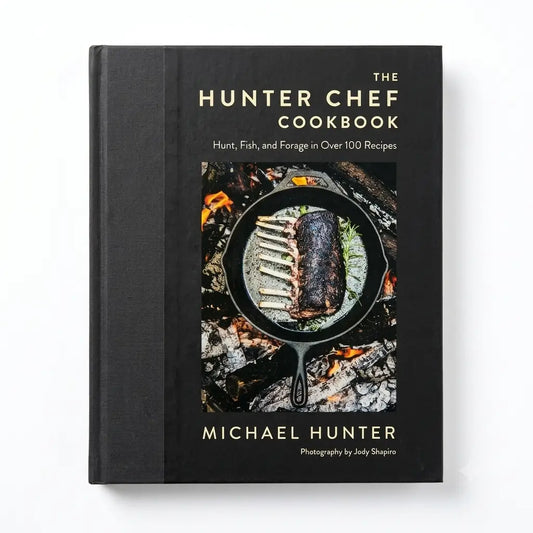 The Hunter Chef Cookbook by Michael Hunter, a hardcover book with a black cover and gold lettering, featuring a central image of ribs cooking in a cast iron skillet over embers.