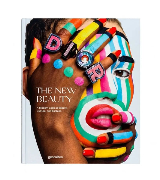 The New Beauty: A Modern Look at Beauty, Culture, and Fashion book cover, featuring a close-up portrait of a person with vibrant multicolor face paint, colorful rings, and bold text on a light gray background.