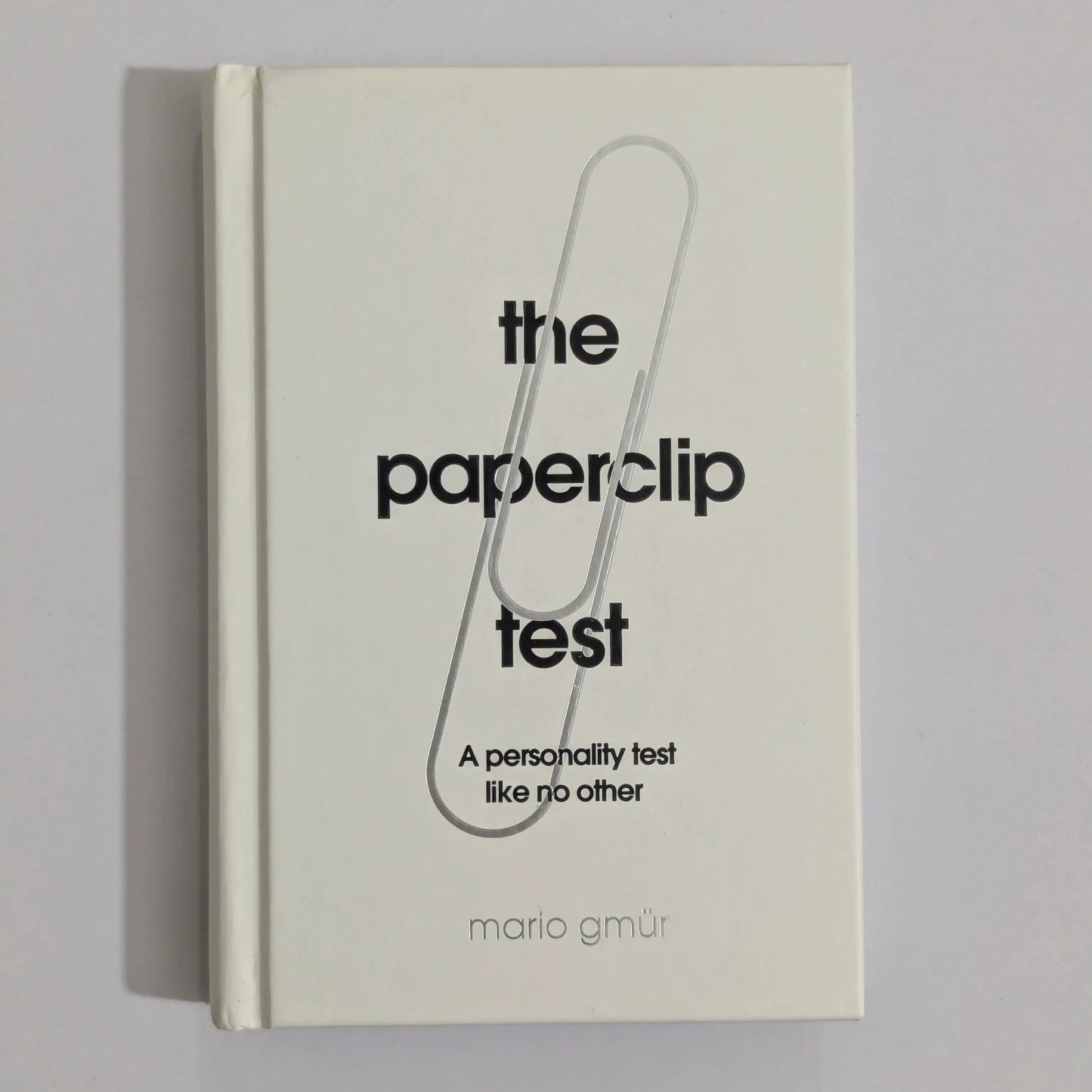 A hardcover book titled 'the paperclip test' by Mario GMUR, featuring a minimalist cream cover with a large black paperclip graphic and black text, photographed against a neutral gray background.
