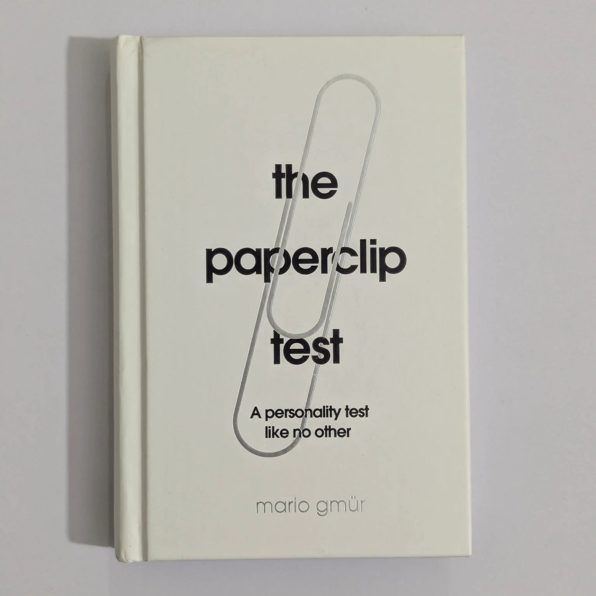 A hardcover book titled 'the paperclip test' by Mario GMUR, featuring a minimalist cream cover with a large black paperclip graphic and black text, photographed against a neutral gray background.