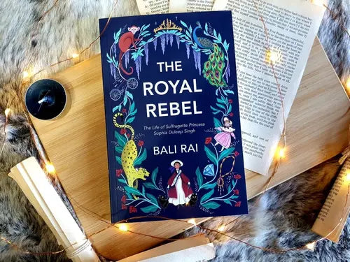 Book titled 'The Royal Rebel' by Bali Rai on a wooden surface with decorative lights and books.