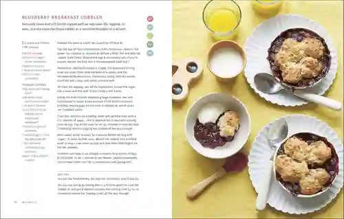 Blueberry breakfast cobbler with a side of ice cream on a yellow tablecloth, next to a recipe book.